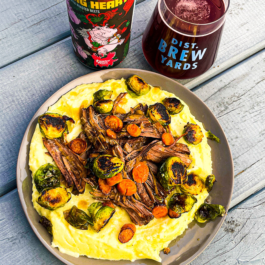 A plate of mashed potatoes, carrots, and brussel sprouts with roasted marks on them, covering a heaping pile of tender chuck roast. A bottle of Bleeding Heart beer from Twisted Hippo is nearby and a poured glass of beer next to it.
