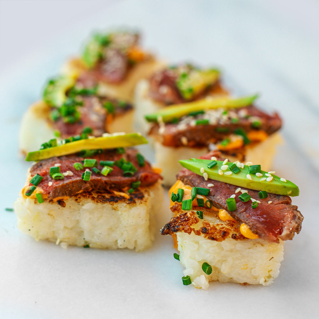 Sushi rice squares with steak on top and avocado and spicy sauce with scallions and sesame seeds