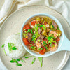 An overhead shot of a bright red gumbo with lots of shrimp and beef and a side of rice and scallions.