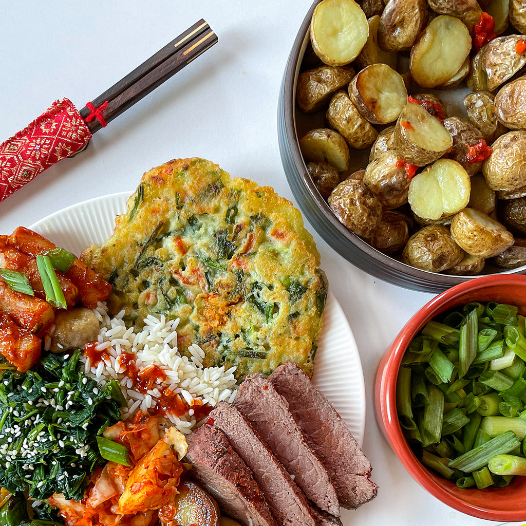 An overhead photo of chopsticks, a big plate of steak and veggies, and a big bowl of potatoes. A small bowl of green onions is on the side.