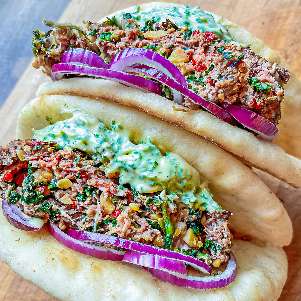 Blended burger patty in a pita with spinach and basil and Mediterranean filling. Sauce and red onions flank each side.