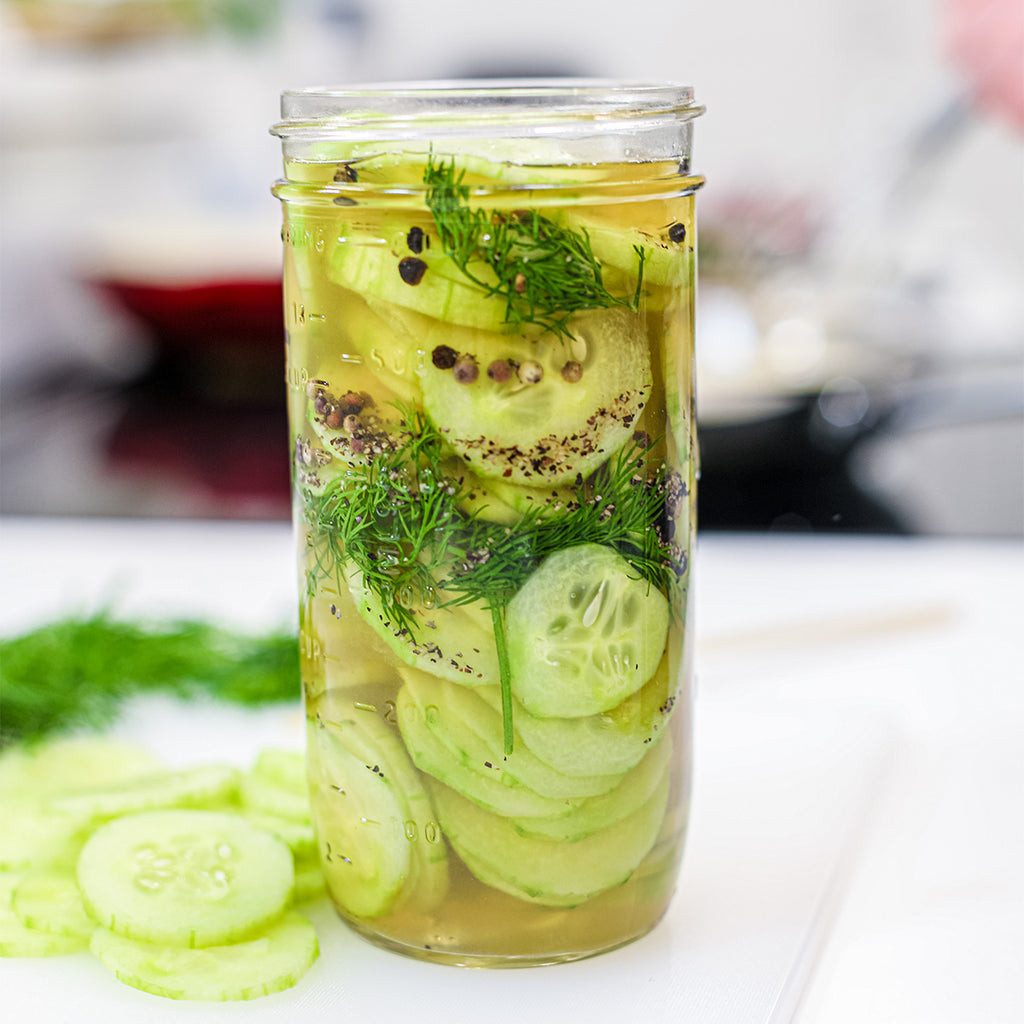A mason jar with fresh dill and cucumbers in brine and spices with a cascade of cucumbers on the cutting board and herbs in the background.
