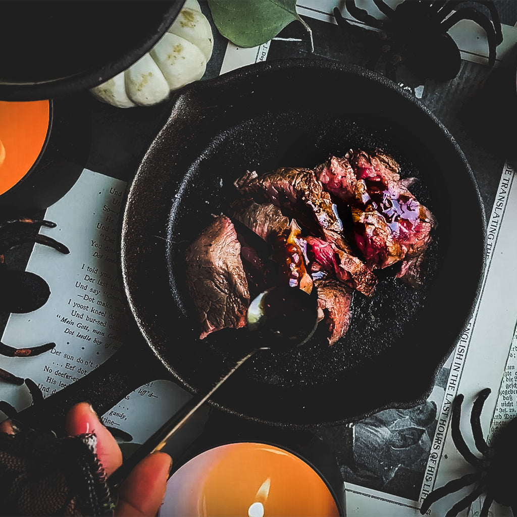 A small cast iron with a filet sliced and someone pouring a red sauce over it with pumpkins and spiders gathered around it.