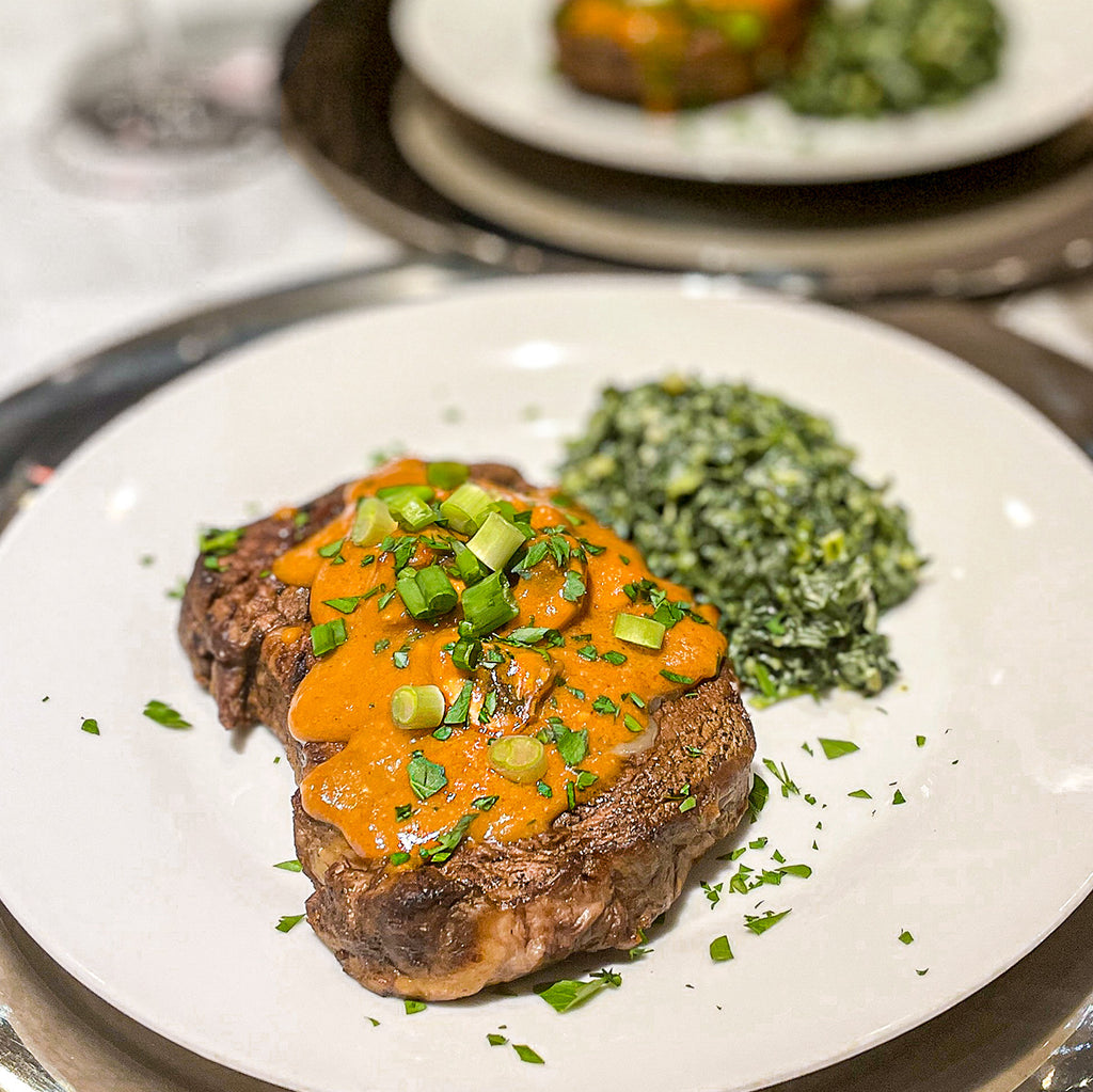 A ribeye steak on a white plate with an orange tinted sauce on top of it and scallions and parsley herbs sprinkled on the sauce. There are greens as a side dish.