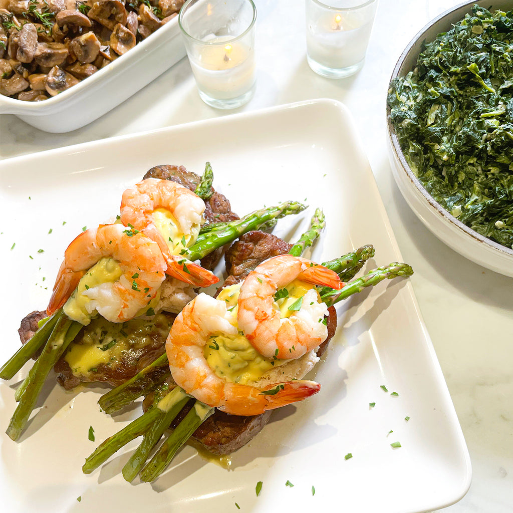 2 steaks with shrimp and a bearnaise sauce on top of them with candles in the background and spinach and mushrooms.