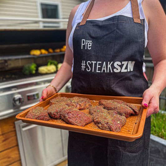 A person holding a tray of steaks with seasoning on them and an apron that has a Pre logo that says #SteakSZN on it