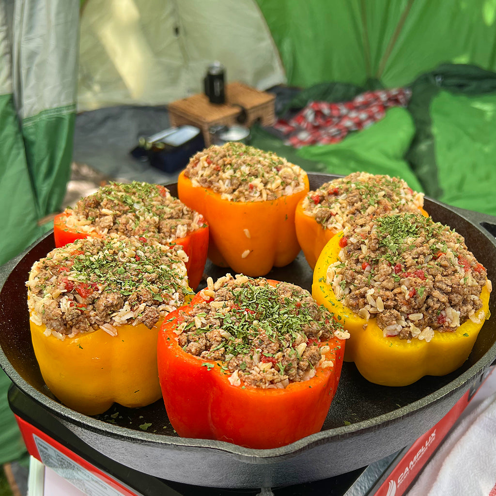 A cast iron pan with red and yellow and orange stuffed peppers with a tent in the background